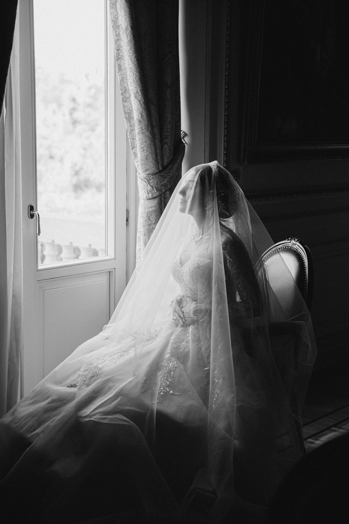 A bride under a flowing veil beside a tall window, in black and white